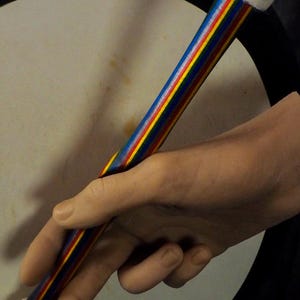 Op de afbeelding: Een kleurrijke drumstok met een blauwe, rode, gele en grijze gestreepte schacht en witte vilten uiteinden. De drumstok wordt in een hand gehouden, met een trommel op de achtergrond. Muziekinstrument.