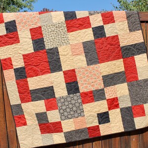 May include: A patchwork quilt with a geometric design. The quilt features squares in red, black, beige, and patterned fabrics. The red squares have a swirling quilted pattern. The quilt is displayed outdoors.