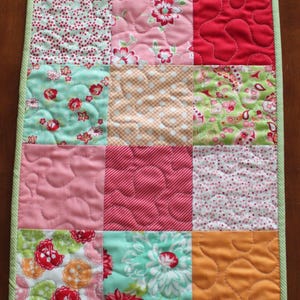 May include: A patchwork table runner featuring a variety of floral and geometric patterns in shades of green, pink, red, and orange. The runner is quilted with a wavy pattern.