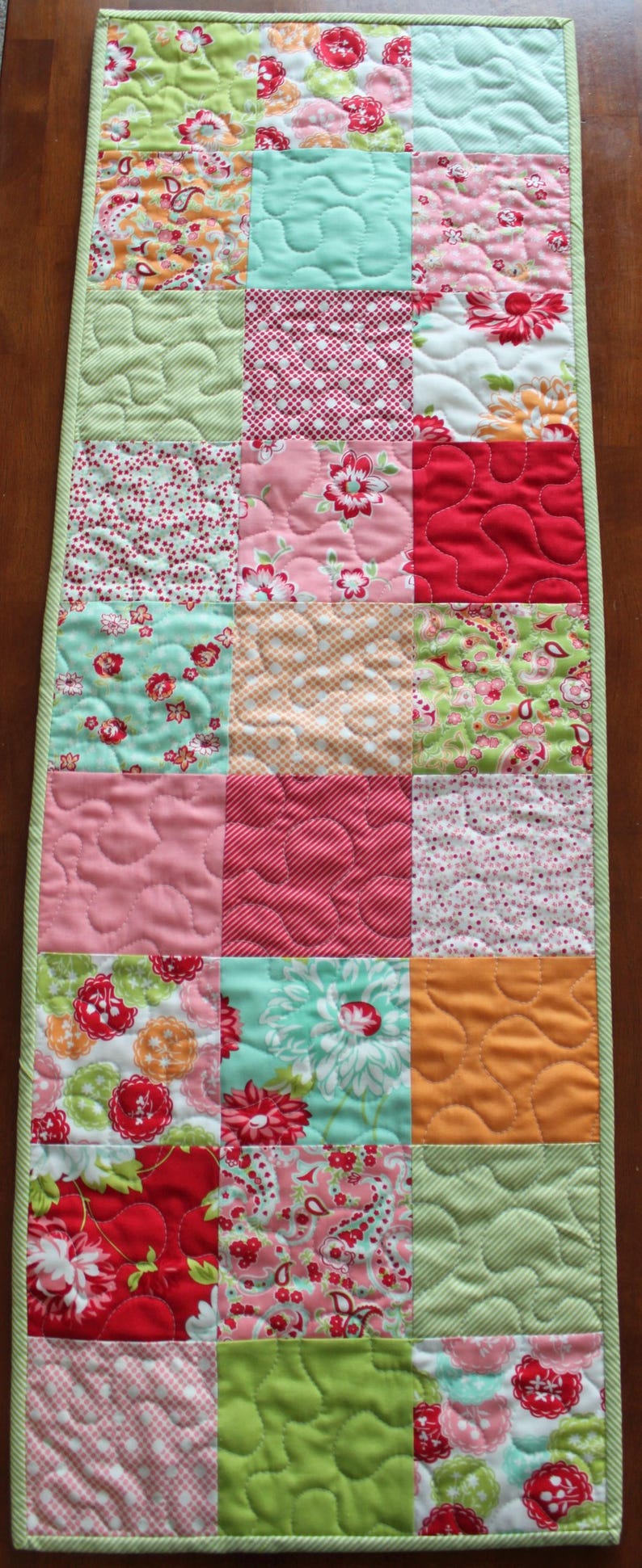 May include: A patchwork table runner featuring a variety of floral and geometric patterns in shades of green, pink, red, and orange. The runner is quilted with a wavy pattern.