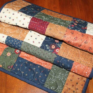 May include: A rectangular quilted table runner featuring a patchwork design of various fabric squares in shades of navy blue, olive green, rust orange, and cream. The fabric squares display floral and vine patterns with visible wavy machine quilting.