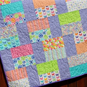 Op de afbeelding: Een patchworkdeken met een paarse achtergrond en kleurrijke vierkanten met verschillende patronen, waaronder vlinders, stippen, strepen en swirls.