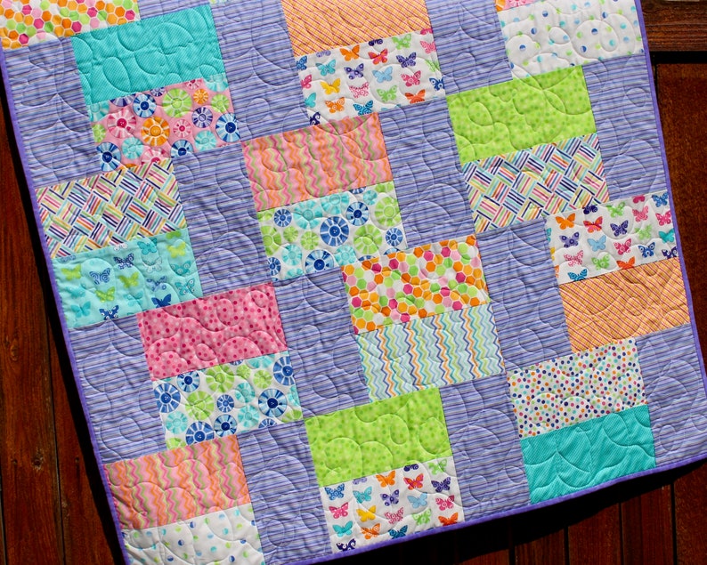 Op de afbeelding: Een patchworkdeken met een paarse achtergrond en kleurrijke vierkanten met verschillende patronen, waaronder vlinders, stippen, strepen en swirls.
