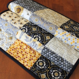 May include: A black and white quilted table runner with a honeycomb pattern, floral design, and a gray and white geometric pattern. The runner is trimmed with black fabric.