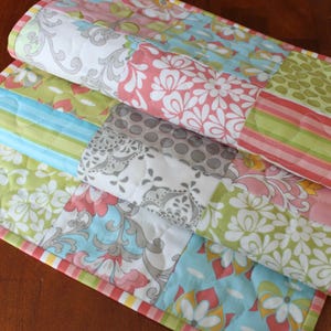May include: A patchwork table runner with a variety of floral patterns in shades of pink, green, blue, and gray. The runner has a yellow, pink, and green border.