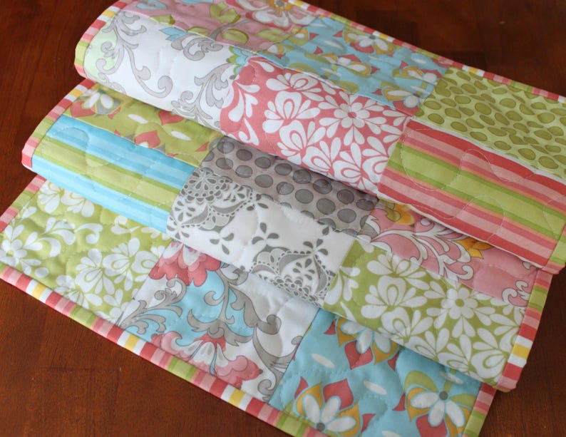 May include: A patchwork table runner with a variety of floral patterns in shades of pink, green, blue, and gray. The runner has a yellow, pink, and green border.