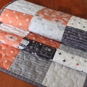 May include: A quilted table runner with a patchwork design featuring orange, black, white, and gray fabrics. The fabrics have different patterns, including geometric shapes, words, and arrows. The runner is folded in thirds, showing the different layers.