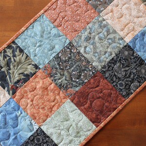 May include: A rectangular quilted table runner featuring a patchwork design of square fabric blocks in shades of burnt orange, dusty blue, sage green, and black with intricate floral and botanical patterns, finished with a solid orange border.