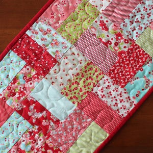 May include: A rectangular quilted table runner featuring a patchwork pattern of small fabric rectangles in shades of red, light blue, white, and lime green. The fabric pieces display various floral and geometric prints, finished with a red border.