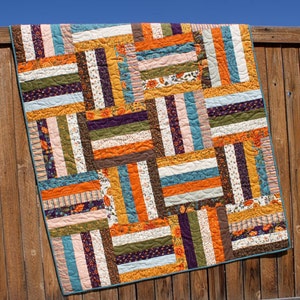 May include: A patchwork quilt with a variety of colorful fabric strips in shades of orange, brown, teal, purple, and white. The quilt is made with a simple pattern of horizontal and vertical strips, creating a geometric design.
