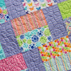 Op de afbeelding: Een kleurrijk patchwork-deken met verschillende patronen, waaronder bloemen, vlinders en geometrische vormen. Het deken is gemaakt van verschillende stoffen in tinten roze, blauw, groen en oranje.