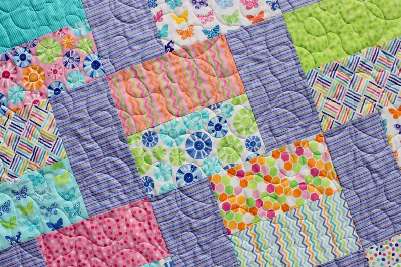 Op de afbeelding: Een kleurrijk patchwork-deken met verschillende patronen, waaronder bloemen, vlinders en geometrische vormen. Het deken is gemaakt van verschillende stoffen in tinten roze, blauw, groen en oranje.