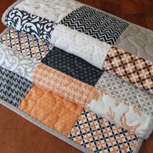 May include: A patchwork quilt with various patterns and colours. The quilt features squares of black and white floral, geometric, and patterned fabrics, along with orange and grey accents. The quilt is bordered with a light grey fabric.