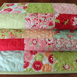 May include: A patchwork quilt table runner featuring a green, red, pink and white floral design. The runner is made from various fabrics and has a quilted texture.