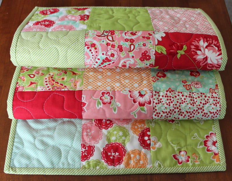 May include: A patchwork quilt table runner featuring a green, red, pink and white floral design. The runner is made from various fabrics and has a quilted texture.