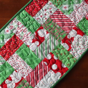 May include: Christmas-themed table runner with a patchwork design. The runner features red, green, and white squares with patterns like snowmen, candy canes, and polka dots. The edges are finished with a green border.