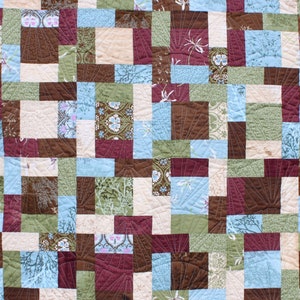 May include: A quilt made of various fabric squares in shades of brown, green, blue, and cream. The squares are arranged in a patchwork pattern, creating a visually interesting and textured design.