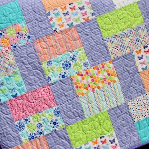 Op de afbeelding: Een kleurrijk patchwork-deken met verschillende patronen, waaronder vlinders, bloemen, stippen en strepen. Het deken is gemaakt van lichtpaarse stof en heeft een gestikt ontwerp.