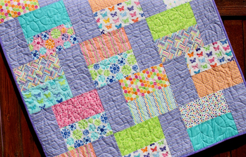Op de afbeelding: Een kleurrijk patchwork-deken met verschillende patronen, waaronder vlinders, bloemen, stippen en strepen. Het deken is gemaakt van lichtpaarse stof en heeft een gestikt ontwerp.