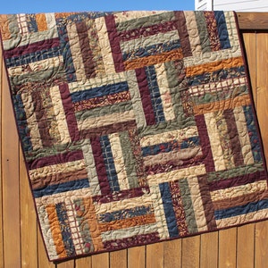 May include: A quilt made of various fabric scraps in shades of brown, green, blue, and orange. The quilt is made in a log cabin pattern with curved lines sewn into the fabric.