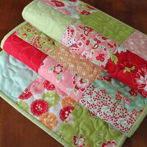 May include: A patchwork quilt runner featuring a mix of floral and geometric patterns in shades of pink, red, green, and white. The runner is made from cotton fabric and has a quilted texture.