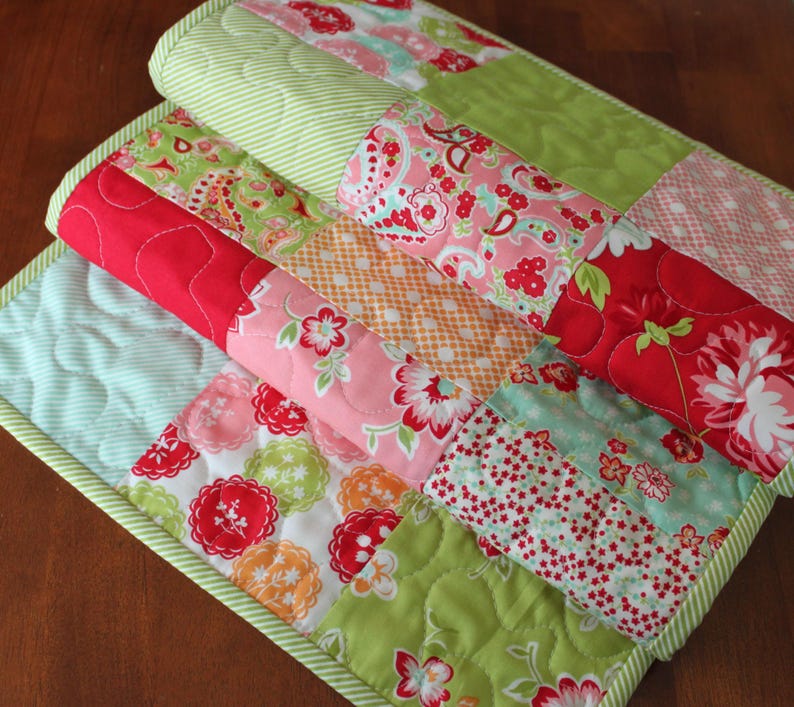 May include: A patchwork quilt runner featuring a mix of floral and geometric patterns in shades of pink, red, green, and white. The runner is made from cotton fabric and has a quilted texture.