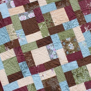 May include: A quilt made of various fabric squares in shades of brown, green, blue, and cream. The squares are arranged in a pattern and quilted with a decorative stitch.
