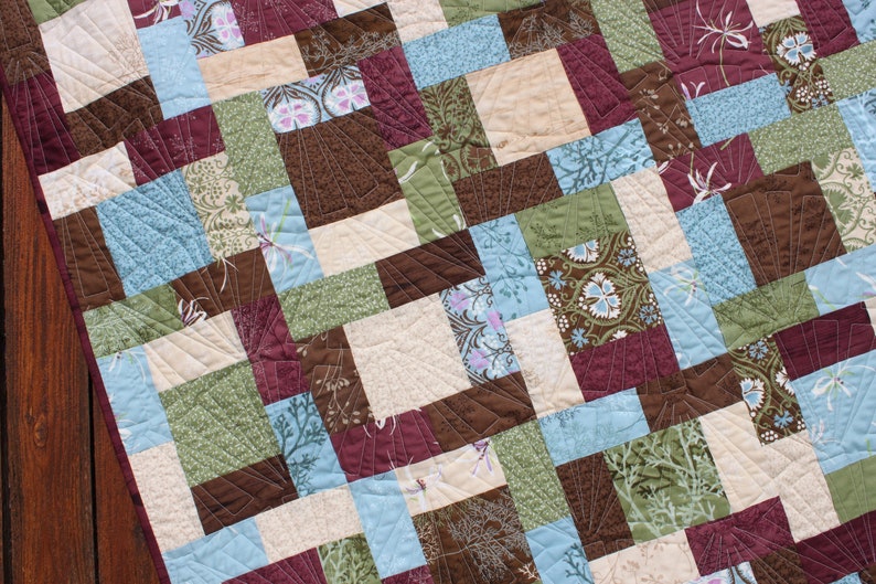 May include: A quilt made of various fabric squares in shades of brown, green, blue, and cream. The squares are arranged in a pattern and quilted with a decorative stitch.