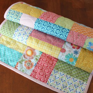 May include: A stack of colourful patchwork quilts with a variety of patterns and colours, including floral, striped, and geometric designs. The quilts have a light pink border and are folded on a wooden surface.