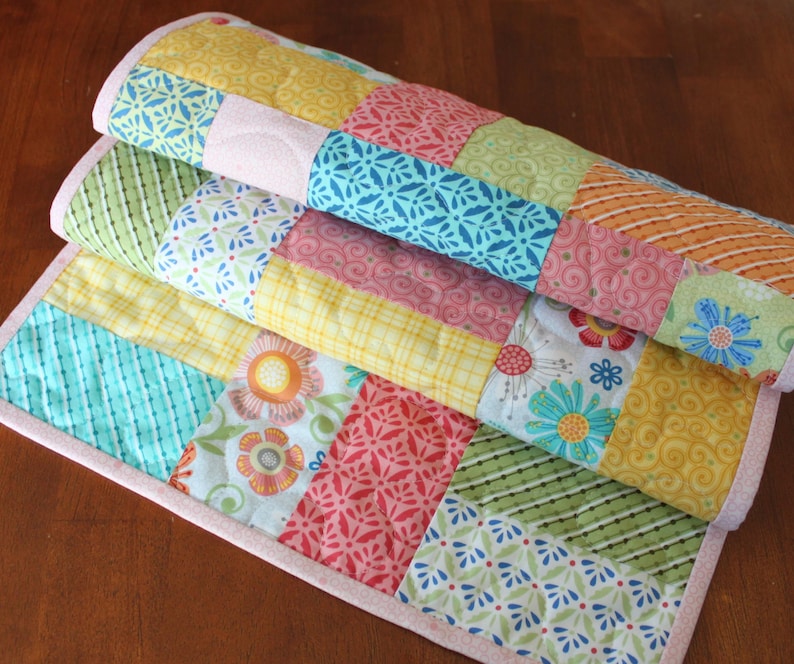 May include: A stack of colourful patchwork quilts with a variety of patterns and colours, including floral, striped, and geometric designs. The quilts have a light pink border and are folded on a wooden surface.