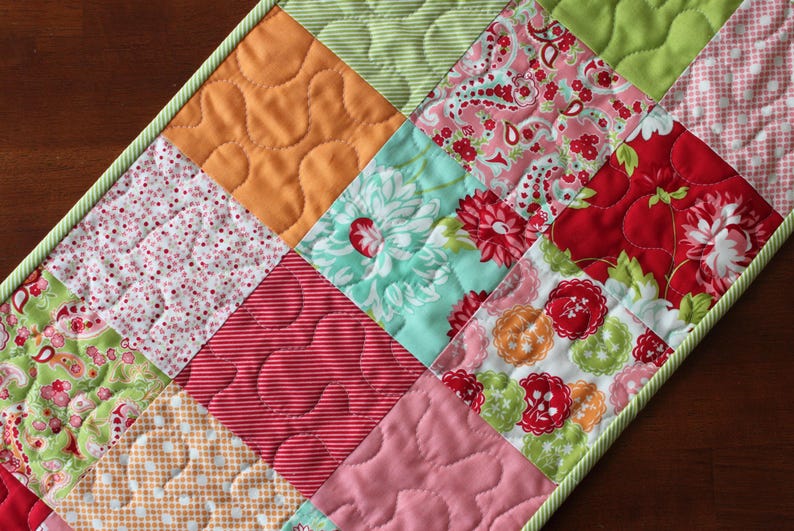 May include: A patchwork table runner featuring a variety of floral and geometric patterns in shades of pink, green, red, and orange. The runner has a light green border.