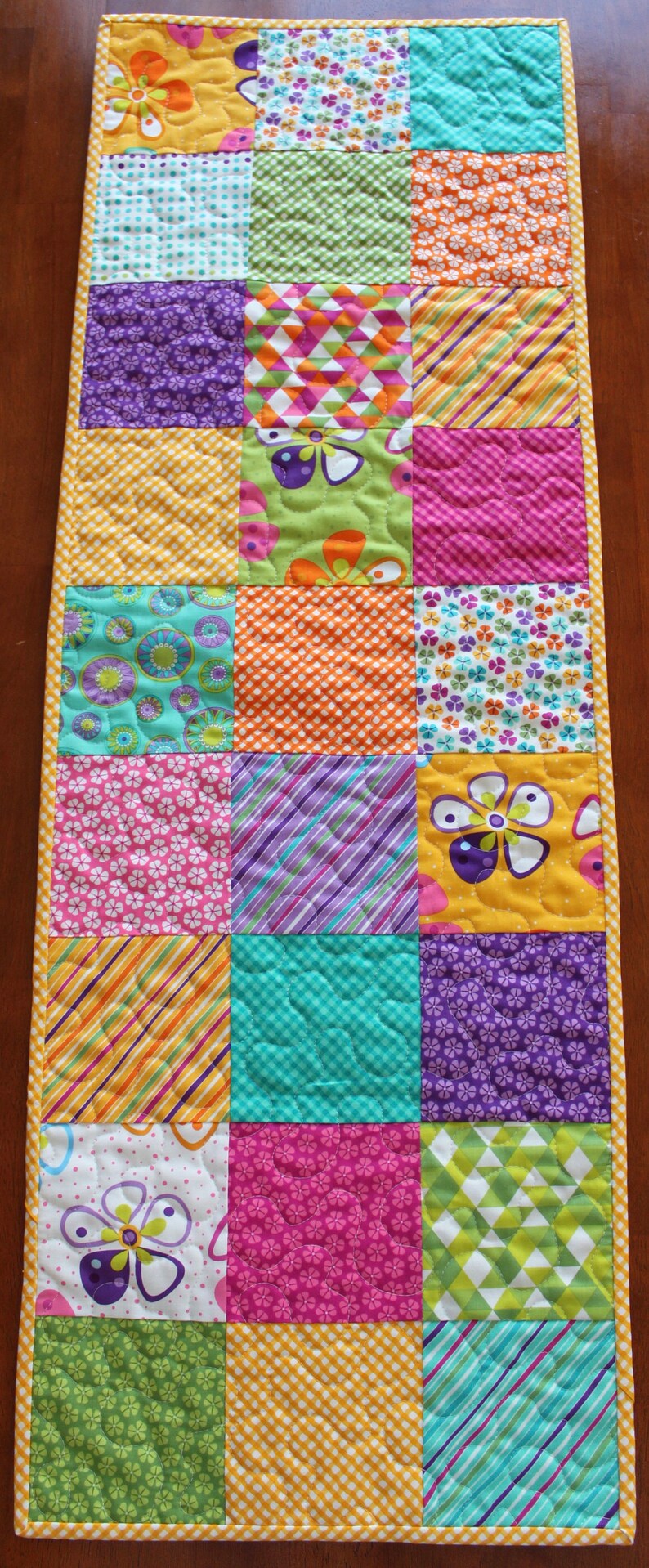 Pu&ograve; includere: Un runner da tavolo patchwork colorato con una variet&agrave; di motivi floreali e geometrici in tonalit&agrave; di giallo, verde, viola, rosa e arancione.