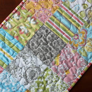 May include: A patchwork table runner with a variety of colorful fabrics, including pink, green, blue, yellow, and gray. The runner is quilted with a heart pattern.