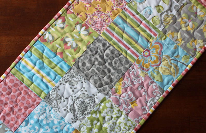 May include: A patchwork table runner with a variety of colorful fabrics, including pink, green, blue, yellow, and gray. The runner is quilted with a heart pattern.