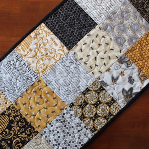 May include: A black and gold table runner with a patchwork design featuring various patterns including bees, honeycomb, and floral motifs. The runner has a black border and the text "Be Positive Believe You Can" is visible.