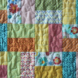 May include: A patchwork quilt with a variety of colourful fabric squares. The squares feature floral, striped, and patterned designs in shades of orange, blue, green, pink, and yellow. The quilt has a quilted texture.