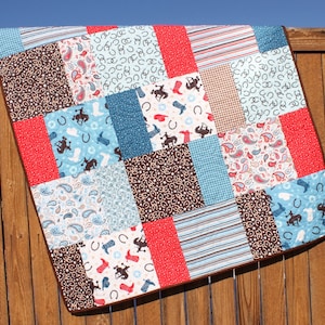 May include: A patchwork quilt with a cowboy theme. The quilt is made of various fabrics in shades of blue, brown, red, and white. The fabrics feature patterns such as paisley, horseshoes, and stars.