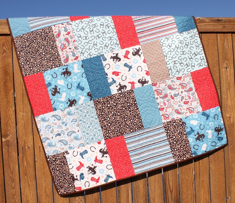 May include: A patchwork quilt with a cowboy theme. The quilt is made of various fabrics in shades of blue, brown, red, and white. The fabrics feature patterns such as paisley, horseshoes, and stars.