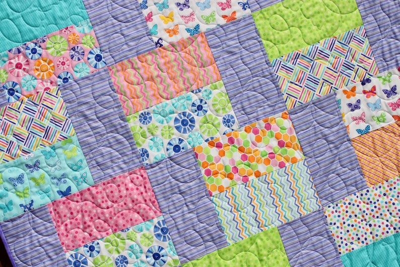 Op de afbeelding: Een kleurrijk patchwork-deken met verschillende patronen, waaronder vlinders, bloemen en geometrische ontwerpen. Het deken is gemaakt van verschillende stoffen in tinten roze, blauw, groen en geel.
