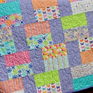 Op de afbeelding: Een patchworkdeken met een paarse achtergrond en kleurrijke vierkanten met verschillende patronen, waaronder vlinders, bloemen en stippen.
