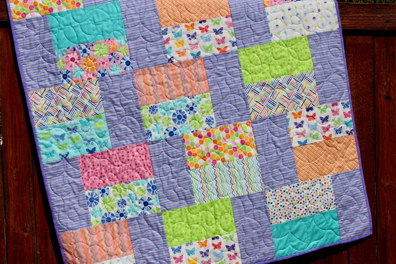 Op de afbeelding: Een patchworkdeken met een paarse achtergrond en kleurrijke vierkanten met verschillende patronen, waaronder vlinders, bloemen en stippen.