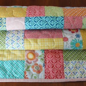 May include: Three colourful patchwork quilts stacked on top of each other. Each quilt has squares with patterns in yellow, pink, blue, green, and orange. The quilts are edged with a pink border.
