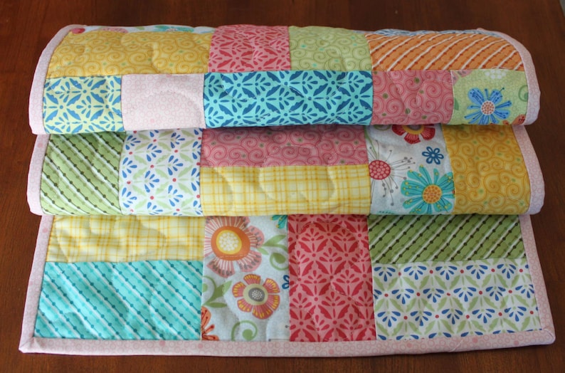 May include: Three colourful patchwork quilts stacked on top of each other. Each quilt has squares with patterns in yellow, pink, blue, green, and orange. The quilts are edged with a pink border.