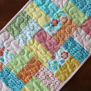 May include: A colorful quilted table runner with a patchwork design. The runner features various patterned fabrics in shades of pink, green, yellow, orange, and blue. Floral and geometric patterns are visible. The runner is approximately 12 inches wide.