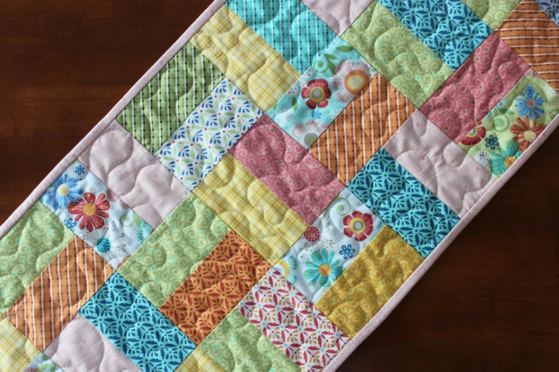 May include: A colourful quilted table runner with a patchwork design. The runner features various patterned fabrics in shades of pink, green, yellow, orange, and blue. Floral and geometric patterns are visible. The runner is approximately 30 cm wide.