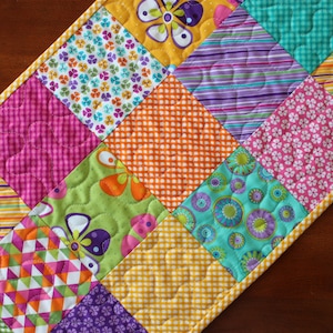 Pu&ograve; includere: Un runner per trapunta patchwork colorato con una variet&agrave; di motivi floreali e geometrici in tonalit&agrave; di rosa, verde, blu, giallo e viola.
