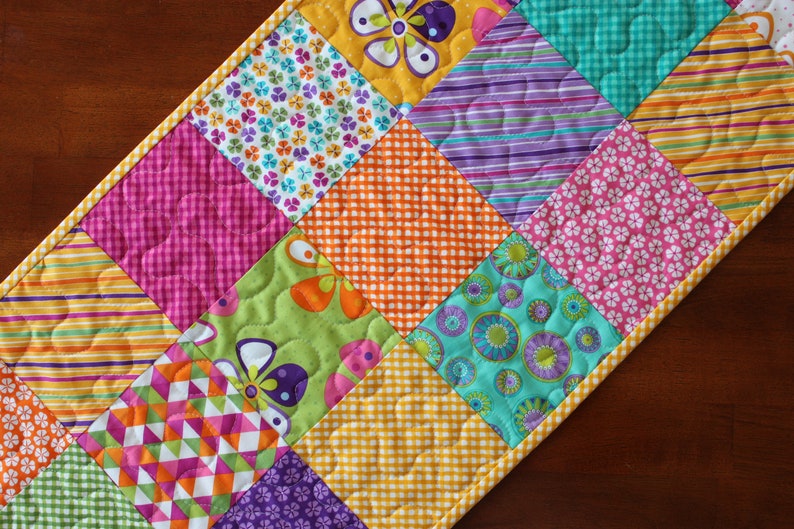 Pu&ograve; includere: Un runner per trapunta patchwork colorato con una variet&agrave; di motivi floreali e geometrici in tonalit&agrave; di rosa, verde, blu, giallo e viola.