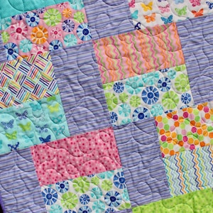 Op de afbeelding: Een paarse en witte quilt met een patchwork-ontwerp met verschillende kleurrijke patronen, waaronder vlinders, bloemen, stippen en strepen.