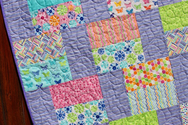 Op de afbeelding: Een paarse en witte quilt met een patchwork-ontwerp met verschillende kleurrijke patronen, waaronder vlinders, bloemen, stippen en strepen.