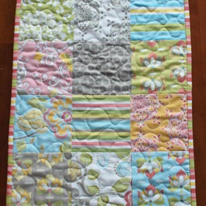 May include: A patchwork table runner with a variety of floral and geometric patterns in shades of pink, green, blue, and gray. The runner has a striped border.